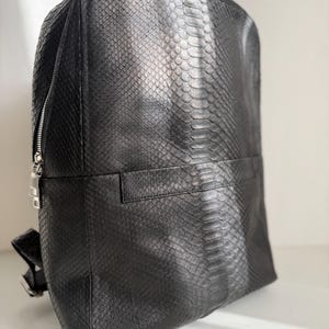 May include: A black backpack featuring a textured snakeskin pattern across the entire exterior. The bag includes a silver-toned metal zipper closure along the side and a horizontal seam detail across the front panel with adjustable straps.