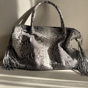 May include: A handbag featuring a grey and white snakeskin pattern with a textured scale finish. The bag includes two top handles and long, dangling tassels on both sides. The design is structured with a central panel and side gussets.