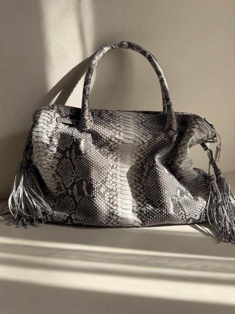 May include: A handbag featuring a grey and white snakeskin pattern with a textured scale finish. The bag includes two top handles and long, dangling tassels on both sides. The design is structured with a central panel and side gussets.