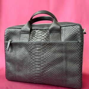 May include: A black briefcase featuring a textured snakeskin pattern across the entire exterior. The bag includes two top handles, a horizontal zippered front pocket with a matching pull tab, and a main zippered compartment with silver hardware.