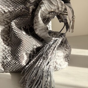 May include: A handbag featuring a textured snakeskin pattern in shades of gray, white, and black. The bag includes a prominent fringe tassel made of the same material, a silver-toned metal zipper closure, and structured handles at the top.