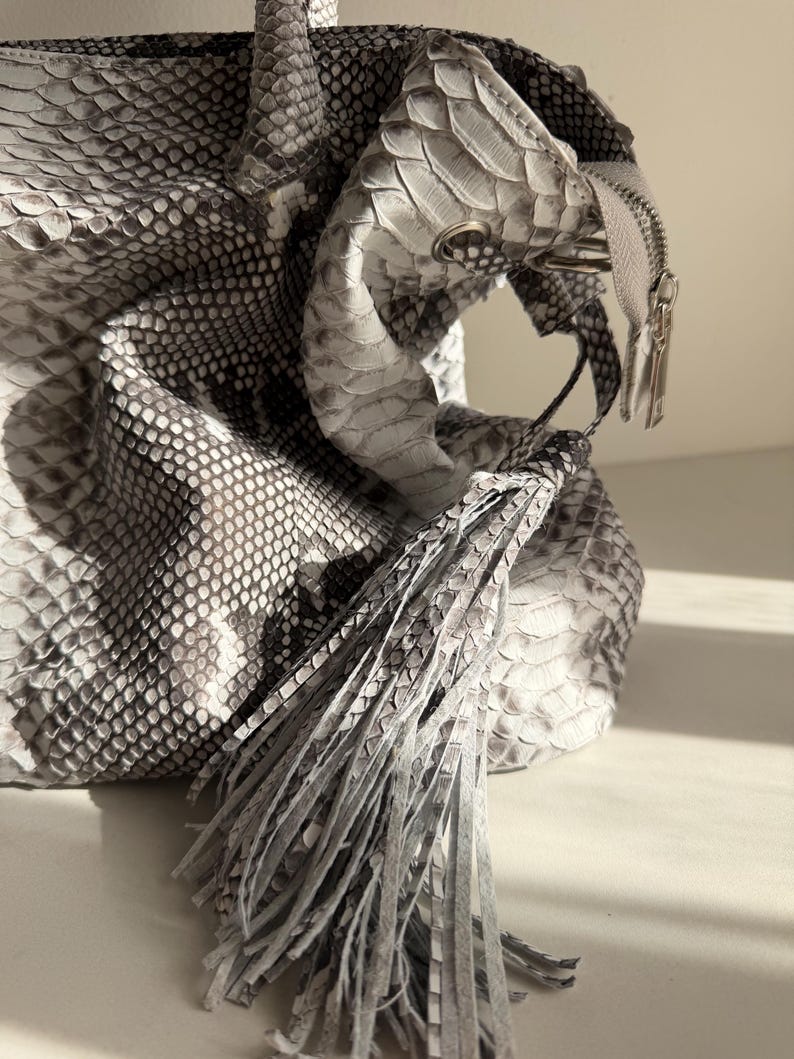 May include: A handbag featuring a textured snakeskin pattern in shades of gray, white, and black. The bag includes a prominent fringe tassel made of the same material, a silver-toned metal zipper closure, and structured handles at the top.