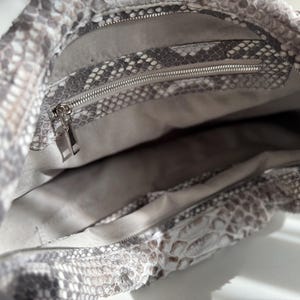 May include: A hobo bag featuring a grey and white snakeskin pattern on the exterior. The interior is lined with a light grey fabric and includes a zippered pocket with a silver-toned metal zipper pull and a separate open compartment.