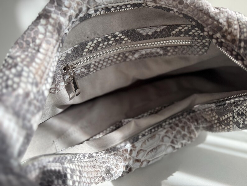 May include: A hobo bag featuring a grey and white snakeskin pattern on the exterior. The interior is lined with a light grey fabric and includes a zippered pocket with a silver-toned metal zipper pull and a separate open compartment.