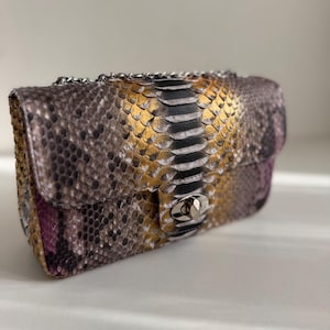Peut inclure: Un sac à bandoulière rectangulaire avec un motif texturé en peau de serpent dans des tons or, noir, gris et violet. Le sac comprend une bandoulière en chaîne métallique argentée et un fermoir rotatif argenté sur le rabat avant.