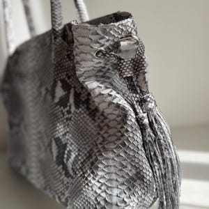 May include: A tote bag featuring a grey and white snakeskin pattern with a metallic silver finish. The bag includes two top handles, a drawstring closure with silver-toned metal eyelets, and a matching snakeskin tassel hanging from the side.