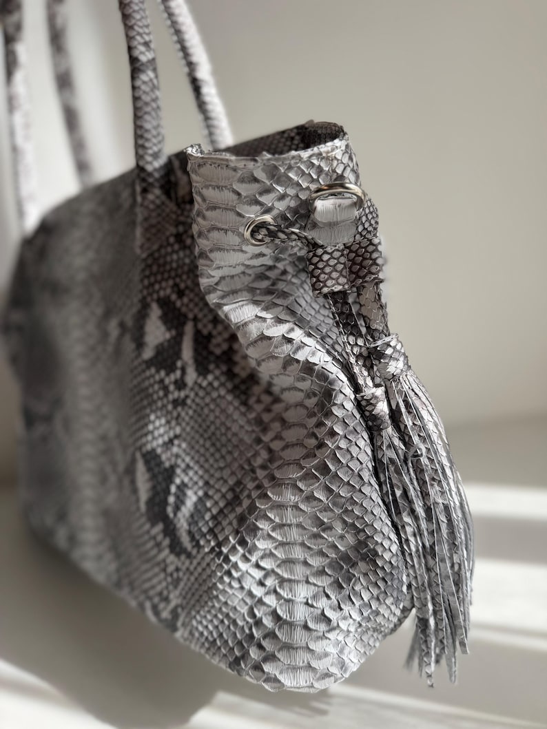 May include: A tote bag featuring a grey and white snakeskin pattern with a metallic silver finish. The bag includes two top handles, a drawstring closure with silver-toned metal eyelets, and a matching snakeskin tassel hanging from the side.