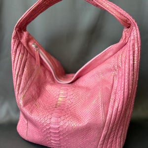 May include: A hobo-style handbag featuring a textured pink snakeskin pattern with subtle gold-toned highlights. The bag has a single integrated shoulder strap, a slouchy silhouette, and a silver-colored zipper closure along the top opening.