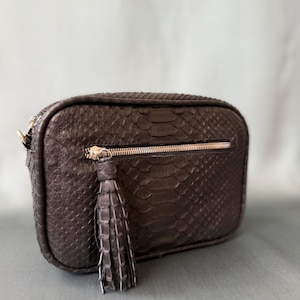 Puede incluir: Un bolso bandolera rectangular de color marrón oscuro con un patrón texturizado de piel de serpiente. El bolso incluye un bolsillo horizontal con cremallera en la parte delantera con una borla de cuero a juego.