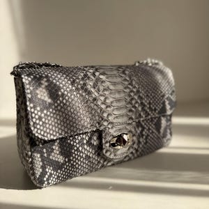 Könnte beinhalten: Eine rechteckige Umhängetasche mit einem grau-schwarzen Schlangenledermuster. Die Tasche verfügt über eine Umschlagklappe mit einem silberfarbenen Drehverschluss aus Metall und einen passenden silberfarbenen Kettenriemen an den oberen Ecken.