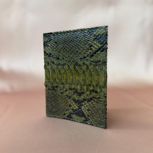 May include: A rectangular card holder featuring a textured, olive green and black snakeskin pattern. The surface displays a detailed, repeating scale design with darker black accents integrated into the natural green tones of the material.