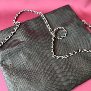 Peut inclure: Un sac à main noir présentant un motif texturé en peau de serpent sur toute la surface. Le sac comprend une longue et fine bandoulière en chaîne métallique argentée, entrelacée d'une fine bande de cuir noir contrastant avec le corps du sac.