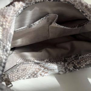 May include: A handbag featuring a textured snakeskin pattern in shades of gray, white, and brown. The interior is lined with a solid light gray fabric and includes two open slip pockets and one zippered pocket along the inner wall.