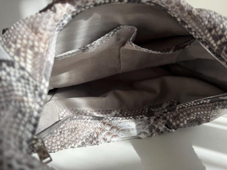 May include: A handbag featuring a textured snakeskin pattern in shades of gray, white, and brown. The interior is lined with a solid light gray fabric and includes two open slip pockets and one zippered pocket along the inner wall.