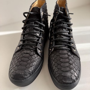 May include: A pair of high-top trainers in black with a textured snakeskin-like pattern. The shoes feature black laces, matching black eyelets, and a solid black rubber sole. The interior lining is a light tan colour, and each shoe has a pull tab.