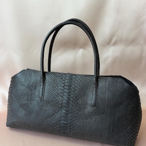 Puede incluir: Un bolso de mano gris carbón oscuro con un patrón texturizado de piel de serpiente en todo el exterior. El bolso presenta una silueta rectangular alargada con dos asas superiores finas y redondeadas a juego en el borde.