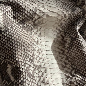 May include: A handbag featuring a textured snakeskin pattern in shades of white, gray, and black. The surface displays a repeating scale design with larger, elongated scales running vertically down the center and smaller scales on the sides.