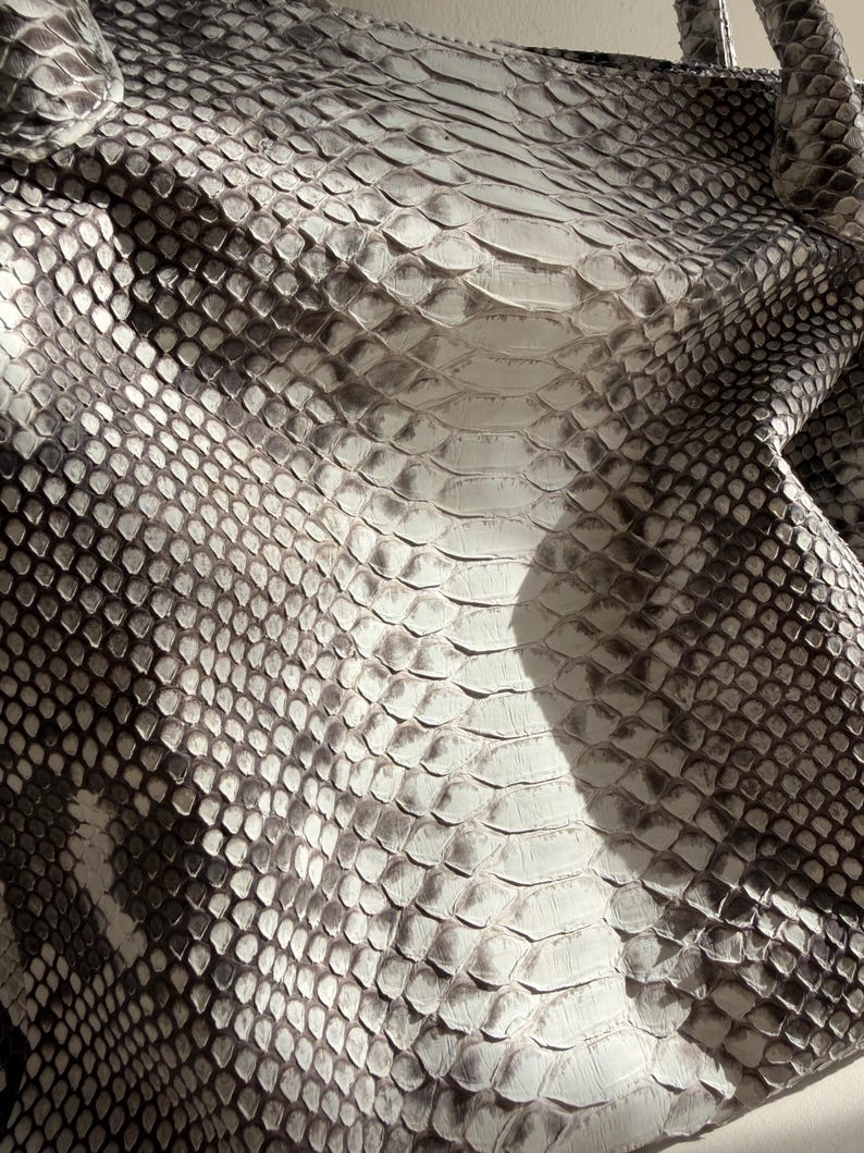 May include: A handbag featuring a textured snakeskin pattern in shades of white, gray, and black. The surface displays a repeating scale design with larger, elongated scales running vertically down the center and smaller scales on the sides.