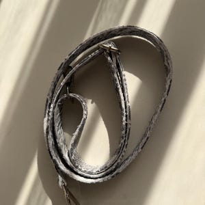 May include: A long, adjustable bag strap featuring a grey and white snakeskin pattern. The strap is constructed from a textured material and includes silver-toned metal hardware, specifically a sliding buckle for length adjustment and a lobster clasp.