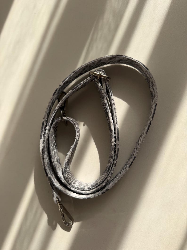 May include: A long, adjustable bag strap featuring a grey and white snakeskin pattern. The strap is constructed from a textured material and includes silver-toned metal hardware, specifically a sliding buckle for length adjustment and a lobster clasp.