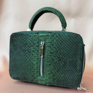 May include: A rectangular dark green handbag with a textured snakeskin pattern. The bag features a single top handle, a vertical silver zipper on the front center, and a wrap-around silver zipper closure along the side edges of the structured body.