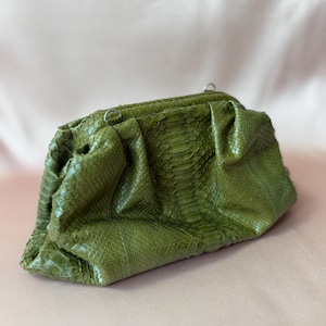 Op de afbeelding: Een gestructureerde olijfgroene clutch met een textuur van slangenleer. De handtas heeft een geplooid ontwerp met een frame-sluiting aan de bovenkant en kleine zilverkleurige metalen ringen aan de bovenrand voor een schouderband.