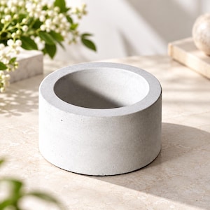 May include: A cylindrical concrete bowl with a smooth, light gray finish and a wide, open top. The vessel features a thick, solid wall construction with a flat base, presenting a neutral, industrial aesthetic suitable for various organizational uses.