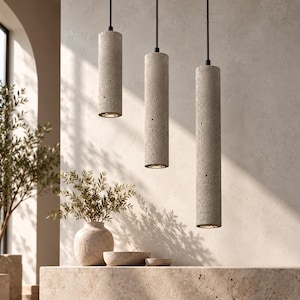 May include: Three cylindrical pendant lights made of grey concrete with a porous, textured surface. Each light hangs from a thin black cord at varying heights, featuring a circular opening at the bottom that emits a soft, downward-facing light.