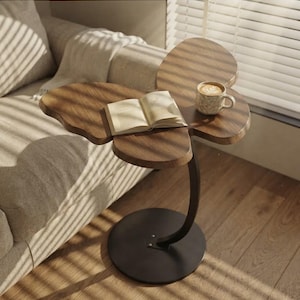 May include: A butterfly-shaped side table with a dark wood grain top, a curved black metal support arm, and a circular black base. An open book and a ceramic mug filled with a coffee beverage are placed on the surface of the wooden tabletop.