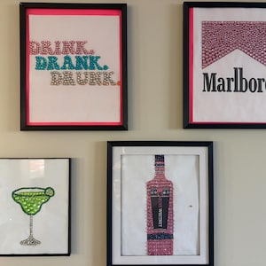 May include: Four framed wall art pieces featuring rhinestone embellishments. One displays the text "DRINK. DRANK. DRUNK." in pink, blue, and silver. Another shows a Marlboro logo with pink rhinestones. A third depicts a green margarita glass, and the fourth shows a pink bottle labeled "PINK WHITNEY."