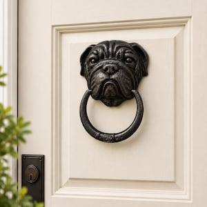 May include: A dark grey, textured metal door knocker shaped like a pug dog head with a circular ring hanging from its mouth. The knocker is mounted on a light cream-coloured panelled door with a black metal lock mechanism visible below it.