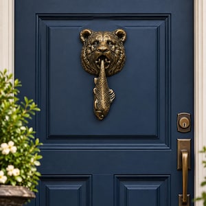Peut inclure: Un heurtoir de porte en métal couleur bronze foncé représentant une tête d'ours tenant un poisson dans sa gueule, fixé sur une porte à panneaux bleu marine avec une poignée et une serrure assorties en bronze sur le côté droit de la porte.