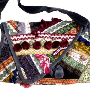 Può includere: Una borsa a tracolla rettangolare con tracolla e bordo in velluto grigio scuro. L'esterno è decorato con un denso collage di strisce di tessuto multicolore, pizzo intricato, nastri intrecciati e una fila di pon pon in velluto rosso scuro.
