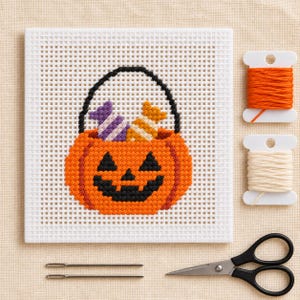 Könnte beinhalten: Ein quadratisches Kunststoffgitter mit einem im Kreuzstich gestickten orangefarbenen Kürbiskorb, der mit violetten und orangefarbenen Bonbons gefüllt ist. Daneben liegen zwei Garnrollen in Orange und Creme, zwei Metallnadeln und eine schwarze Schere.