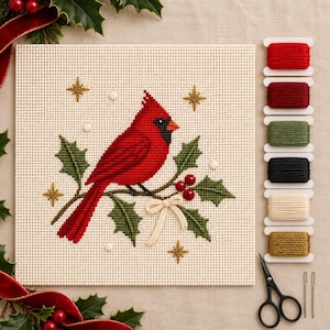May include: A cross-stitch kit featuring a red cardinal bird perched on a holly branch with green leaves, red berries, and a cream bow. The design includes gold star accents on cream fabric. Includes six spools of embroidery thread and scissors.