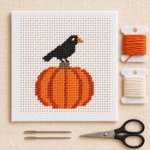 Könnte beinhalten: Ein quadratischer weißer Aida-Stoff mit einem Kreuzstichmotiv, das eine schwarze Krähe auf einem orangefarbenen Kürbis zeigt. Enthalten sind zwei weiße Kunststoffspulen mit orangefarbenem und cremefarbenem Stickgarn, zwei silberne Nadeln und eine schwarze Schere.