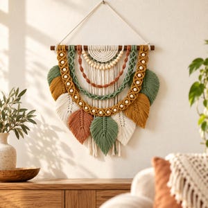 May include: A macrame wall hanging suspended from a wooden dowel, featuring layered cotton cord in shades of sage green, mustard yellow, terracotta, and cream. The design includes intricate knotted arches, wooden beads, and leaf-shaped fringe.