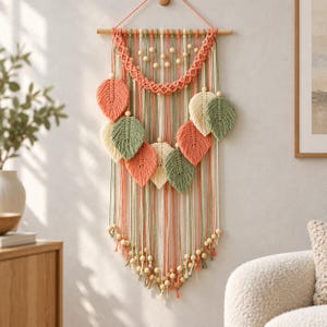 Pode incluir: Uma tapeçaria de macramé com um suporte de madeira, cordões de algodão em coral e verde sálvia, e contas de madeira natural. O design inclui um arco central trançado, vários elementos em forma de folha em coral, sálvia e creme, e franjas com contas.