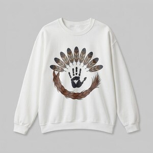 Könnte beinhalten: Ein weißes Langarm-Sweatshirt mit einem zentralen Grafikdesign. Der Aufdruck zeigt einen schwarzen Handabdruck über einem Kreis aus braunem geflochtenem Haar, umgeben von einem Bogen aus zehn braunen und grauen Federn in Fächerform.