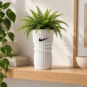 Op de afbeelding: Een witte cilindrische plantenpot met een geribbeld, gestructureerd oppervlak en een zwart Nike swoosh-logo op de voorzijde. De pot bevat een levendige groene varenplant met delicate, vederachtige bladeren die uit de opening naar buiten steken.