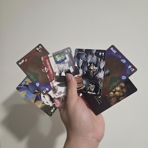Puede incluir: Una mano sosteniendo un abanico de cinco cartas de juego. Las cartas presentan varias ilustraciones, incluyendo un logo de Sprite, un personaje etiquetado como Recruit y otras marcadas como Buff con valores numéricos. Las cartas tienen arte oscuro.