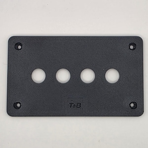 May include: A rectangular black plastic faceplate featuring four circular cutouts arranged in a horizontal row. The plate includes four countersunk screw holes at the corners and the embossed text T&B centred near the bottom edge.