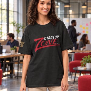 May include: A black short-sleeve t-shirt featuring the text STARTUP ZONE in white and red on the chest. The right sleeve displays a smaller logo with the text STARTUP ZONE ORG. The shirt is paired with light-colored pants.
