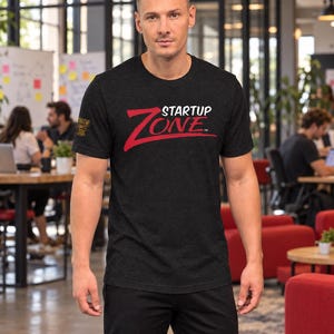 May include: A charcoal gray short-sleeve t-shirt featuring the text STARTUP ZONE in white and red on the chest. A smaller, gold-colored logo is printed on the right sleeve. The shirt has a crew neckline and a standard fit, constructed from a heathered fabric.