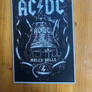 Può includere: Un adesivo rettangolare con sfondo nero e una grafica bianca dall'aspetto usurato raffigurante una grande campana. Il testo AC/DC appare in alto e sulla campana, con le parole HELLS BELLS stampate sotto la campana, circondate da fulmini.