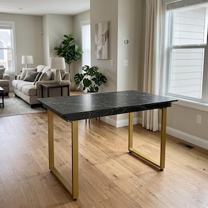 Puede incluir: Una mesa rectangular con una superficie negra y vetas blancas que imitan el mármol. La mesa se apoya sobre cuatro patas robustas de color dorado metálico, dispuestas en dos marcos en forma de U en cada extremo de la estructura.