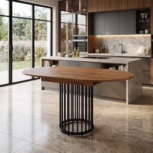 Peut inclure: Une table à manger ovale dotée d'un plateau en bois naturel et d'une base cylindrique composée de tiges métalliques noires verticales. La table est placée dans un espace cuisine avec un îlot en marbre et des armoires sombres en arrière-plan.