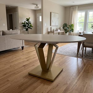 May include: A dining table with an oval-shaped white top and a geometric pedestal base finished in a matte gold color. The base consists of a square floor plate supporting angled metal supports that connect to the underside of the table.