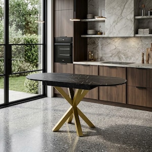 Puede incluir: Una mesa de comedor ovalada con una superficie de mármol negro con vetas blancas y una base geométrica de metal dorado. La mesa se encuentra sobre un suelo gris moteado en una cocina con armarios de madera oscura y un salpicadero de mármol.