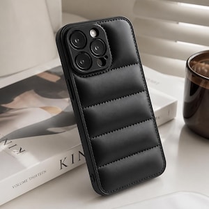 May include: A black smartphone case featuring a padded, quilted design that mimics the appearance of a puffer jacket. The case has a rectangular shape with horizontal stitching and a dedicated cutout section for a triple-lens camera system.